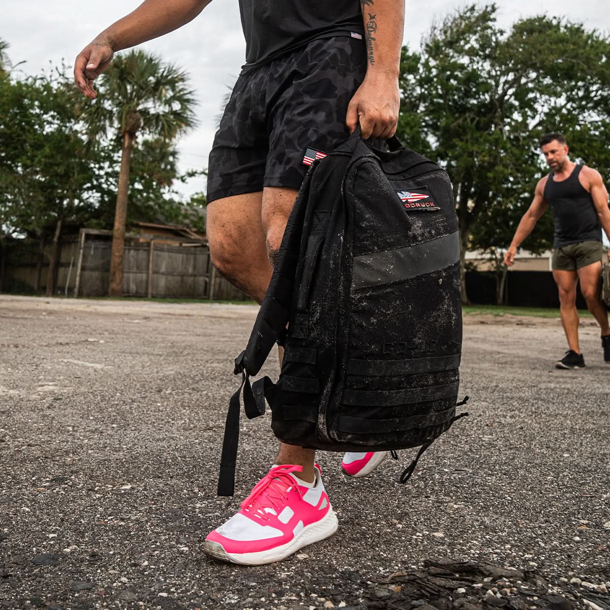 Two men participating in outdoor rucking with backpacks are fully equipped with the GORUCK Ruck Clubs Starter Kit - Light, ensuring enhanced endurance and comfort, while one of them sports bright pink shoes.