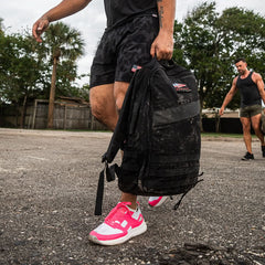 Two men participating in outdoor rucking with backpacks are fully equipped with the GORUCK Ruck Clubs Starter Kit - Light, ensuring enhanced endurance and comfort, while one of them sports bright pink shoes.