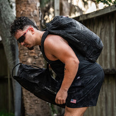 A man sporting sunglasses lifts weights under the open sky, his black Ruck Clubs Starter Kit - Light conveniently accommodating all of GORUCK's features. Clad in dark sportswear, he's prepared for any challenge with a style that effortlessly combines function and form.