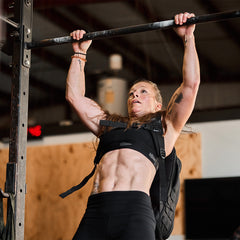 In a bustling gym, a determined individual performs pull-ups on a bar, wearing the Rucker 4.0 from GORUCK to simulate the challenge of rucking. The intensity is palpable as they push their limits in pursuit of strength and endurance.