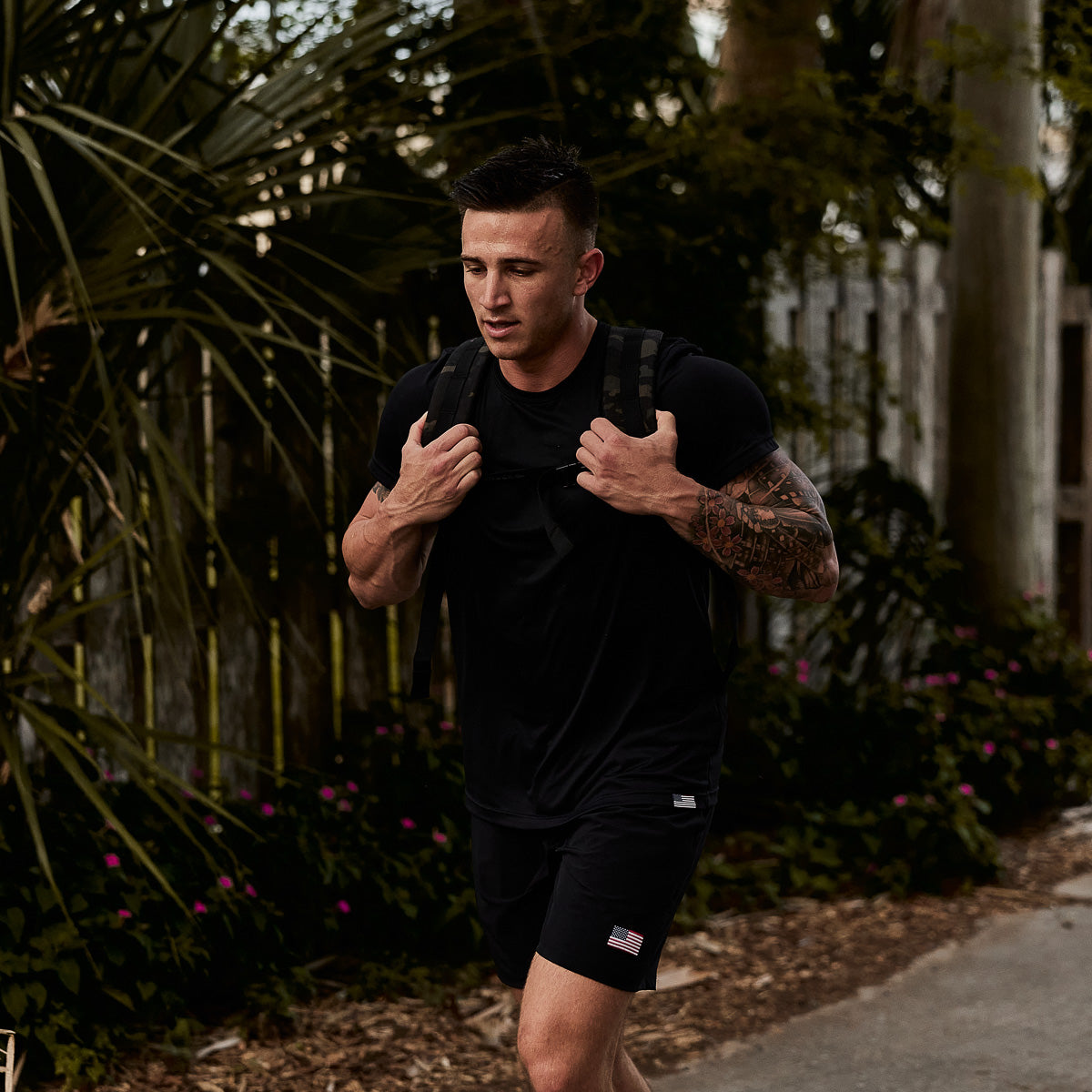 A man in black athletic wear carries the GORUCK Ruck Plate Carrier 3.0, featuring ergonomic lumbar support, as he walks outdoors along a plant-lined path with a wooden fence.