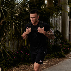 A man in black athletic wear carries the GORUCK Ruck Plate Carrier 3.0, featuring ergonomic lumbar support, as he walks outdoors along a plant-lined path with a wooden fence.