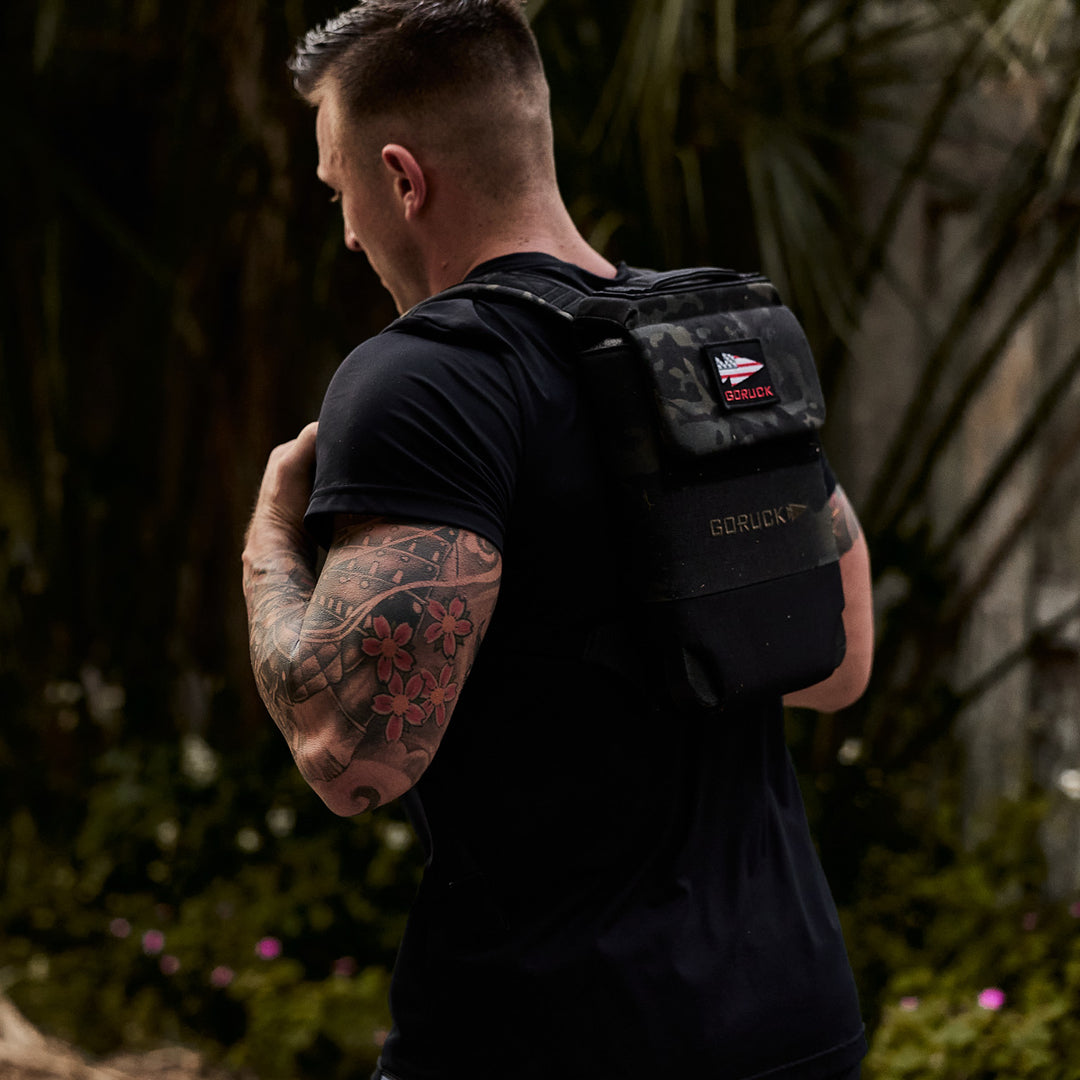 Man with floral arm tattoos wearing a black GORUCK Ruck Plate Carrier 3.0, made with ballistic nylon and ergonomic lumbar support, outdoors and facing away from the camera.