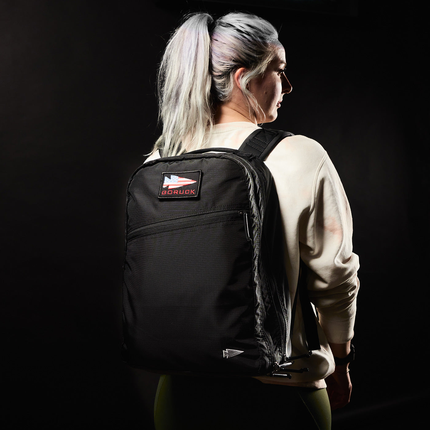 A woman with silver hair wears a black Goruck backpack, standing against a dark background.