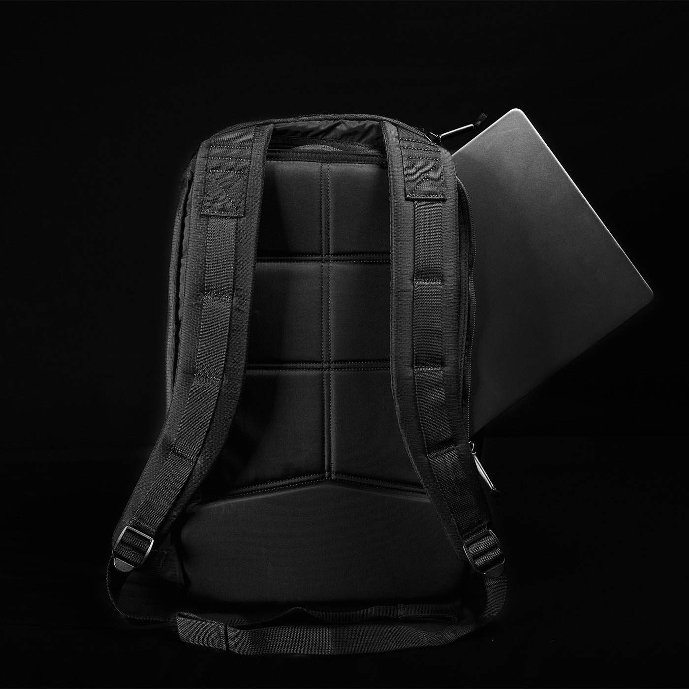 Black backpack with padded straps and a laptop partially visible in the main compartment against a dark background.