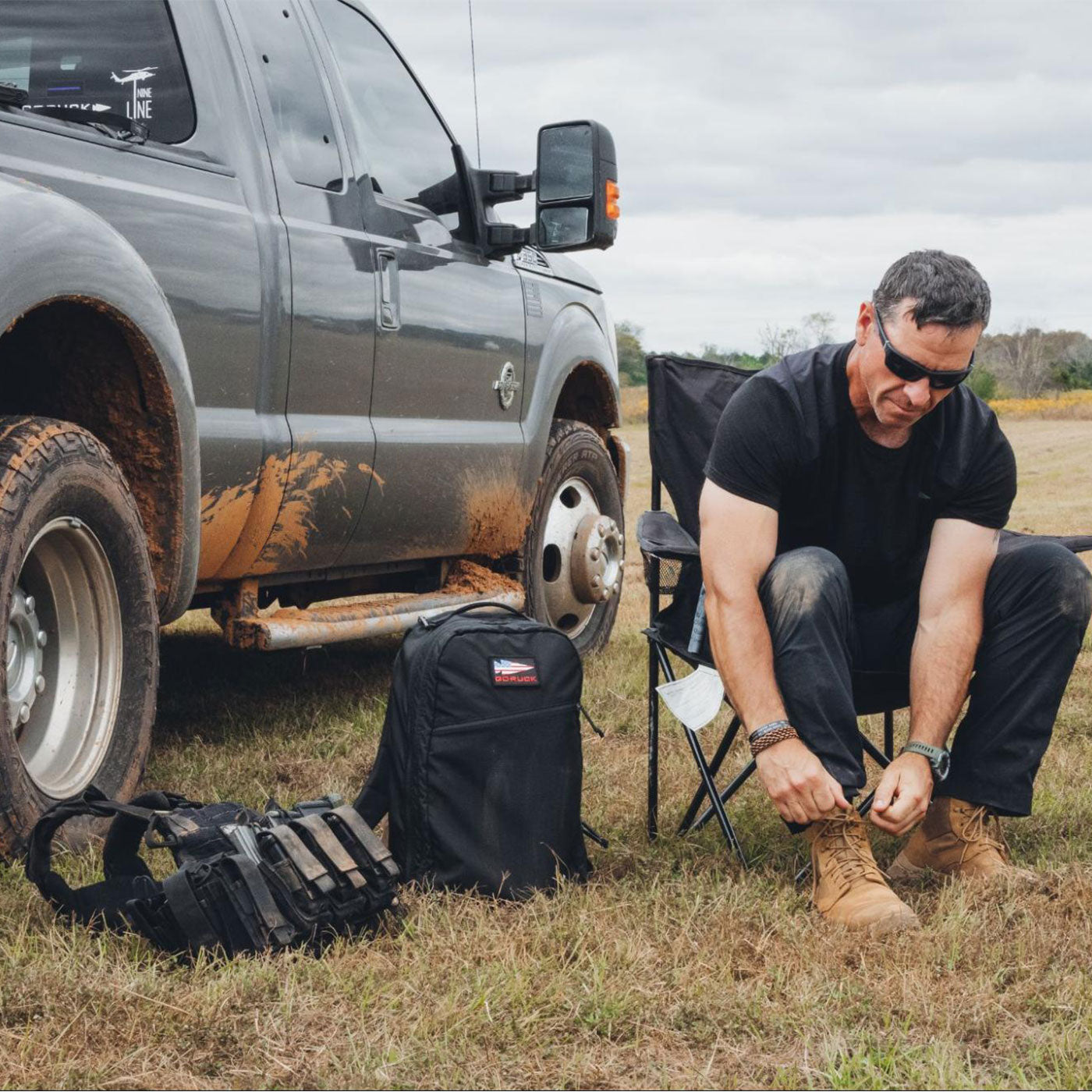 Man sitting by a muddy truck, tying boots, with a backpack and gear on the grass beside him.