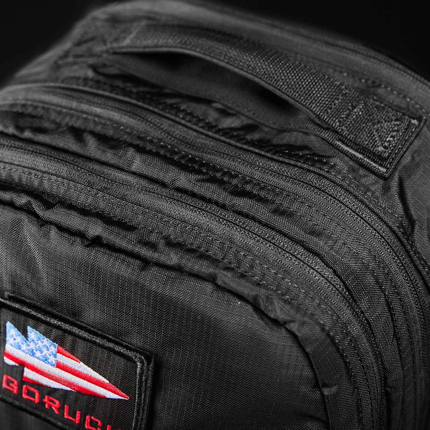 Close-up of a black backpack with an American flag patch and multiple zippered compartments.