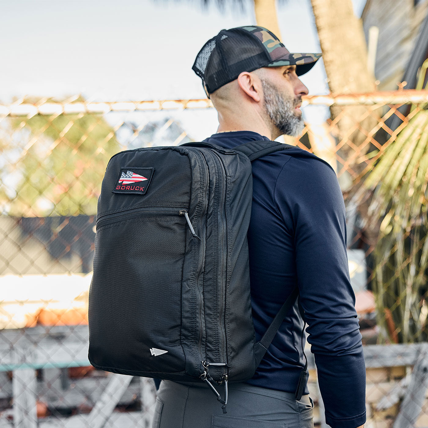 Man wearing a black backpack and a cap, standing outdoors near a chain-link fence with trees in the background.