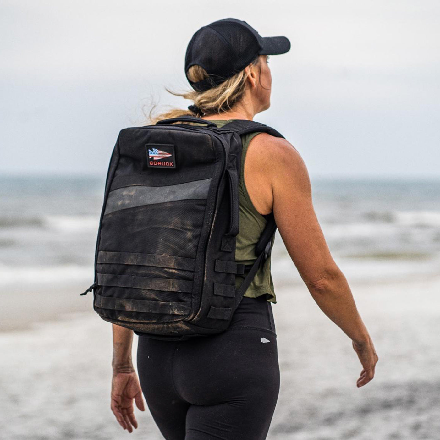 Woman wearing a black backpack walks on a sandy beach, facing the ocean, under a cloudy sky.