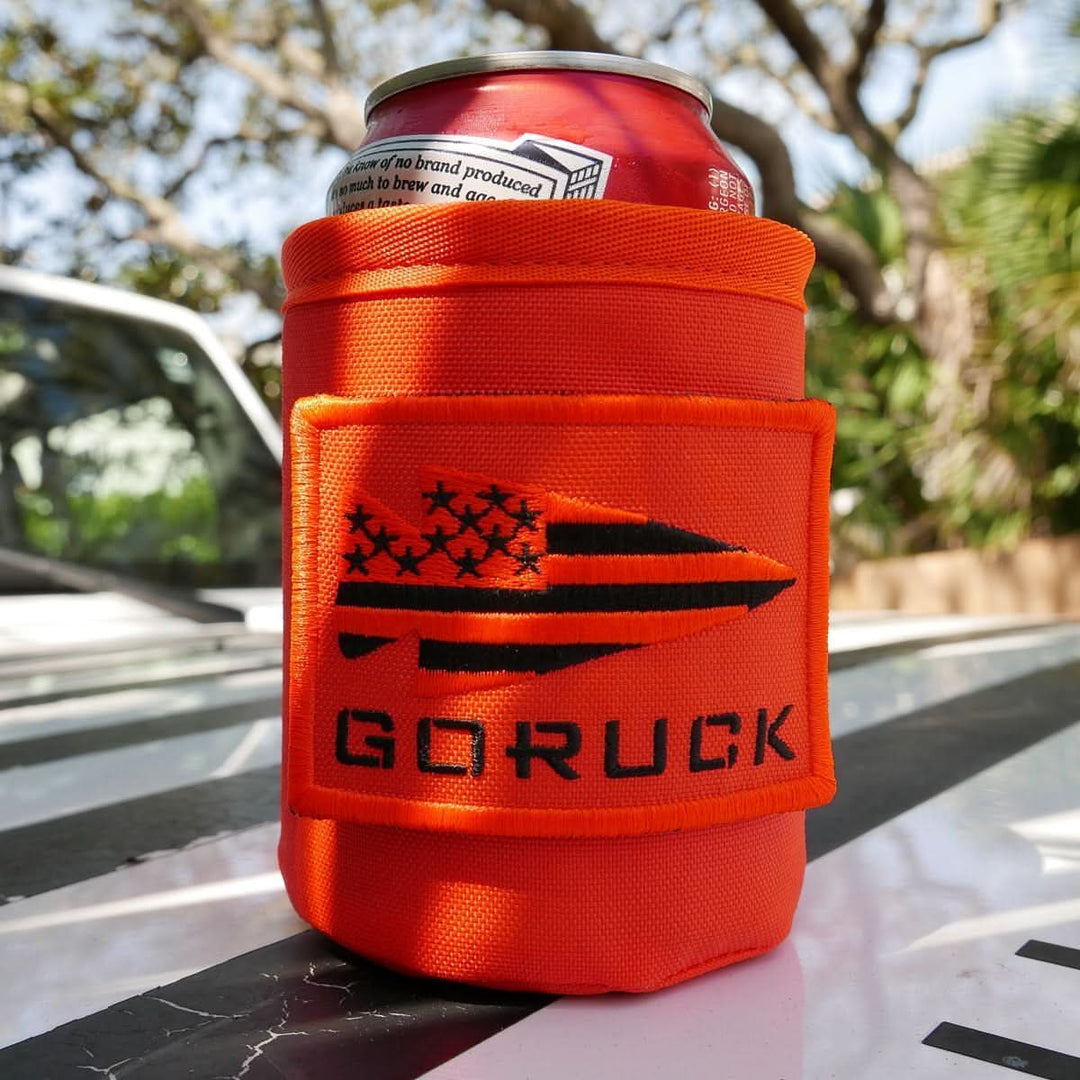 A red can of cold beer sits outdoors on a white surface in a GORUCK Ballistic Beer Jacket, an orange 1000D CORDURA koozie with a black GORUCK logo.