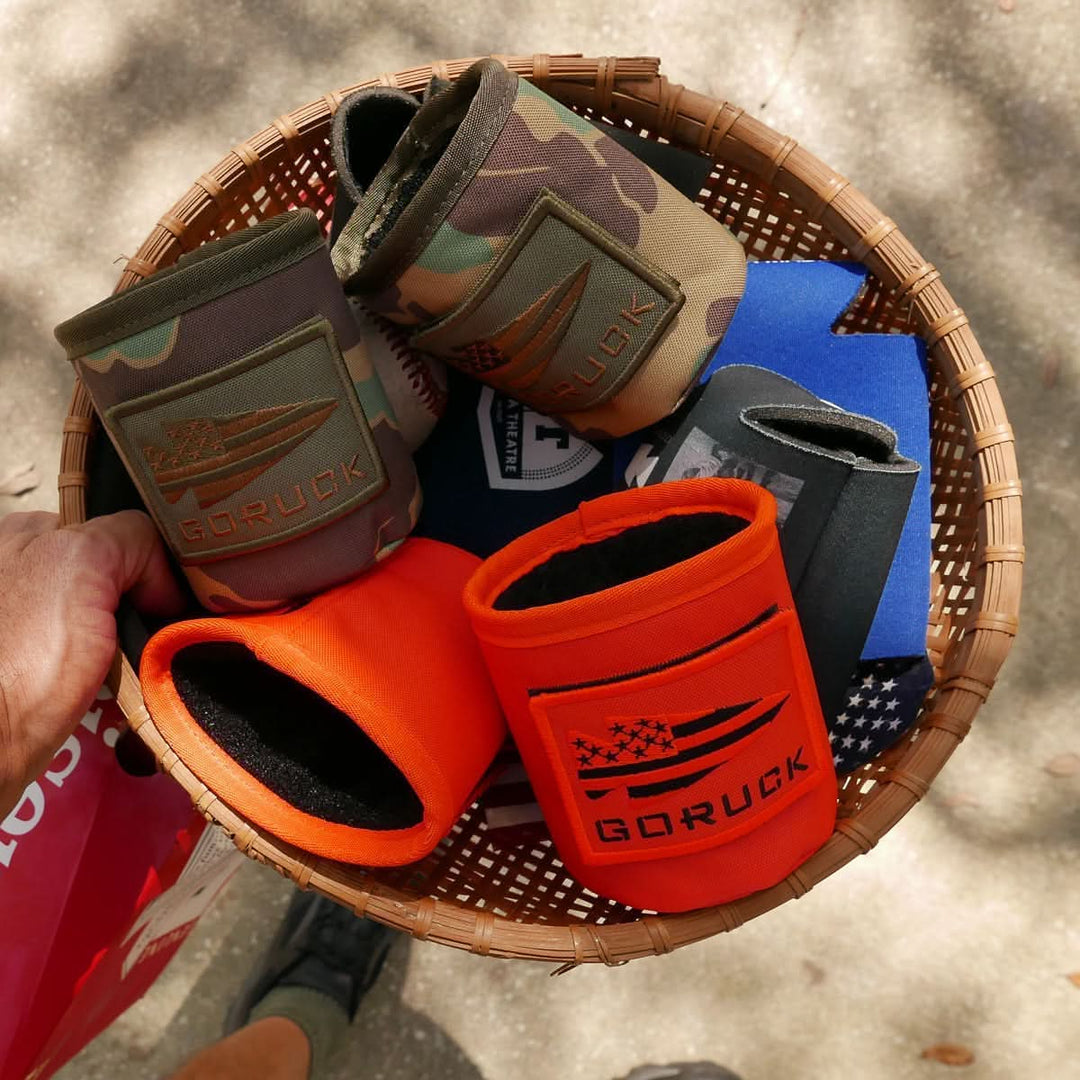 A hand holds a basket of GORUCK Ballistic Beer Jackets, made from rugged 1000D CORDURA in camo and orange—perfect for keeping your cold beer chilled.