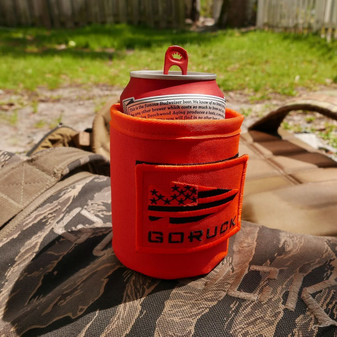 A Budweiser can in the GORUCK Ballistic Beer Jacket, made from rugged 1000D CORDURA, rests on camouflage gear outdoors on a sunny day, ready to keep your beer cold.