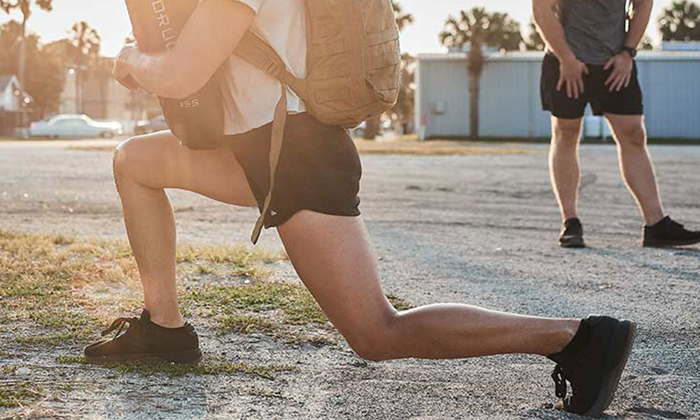 Person lunging outdoors with a backpack, while another stands in the background on a sunny day.