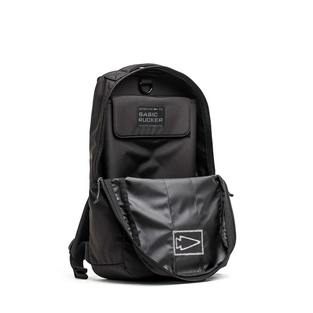 Black Basic Rucker® backpack with an open front pocket, displaying a patch labeled "Basic Rucker®" and a stitched arrow design.