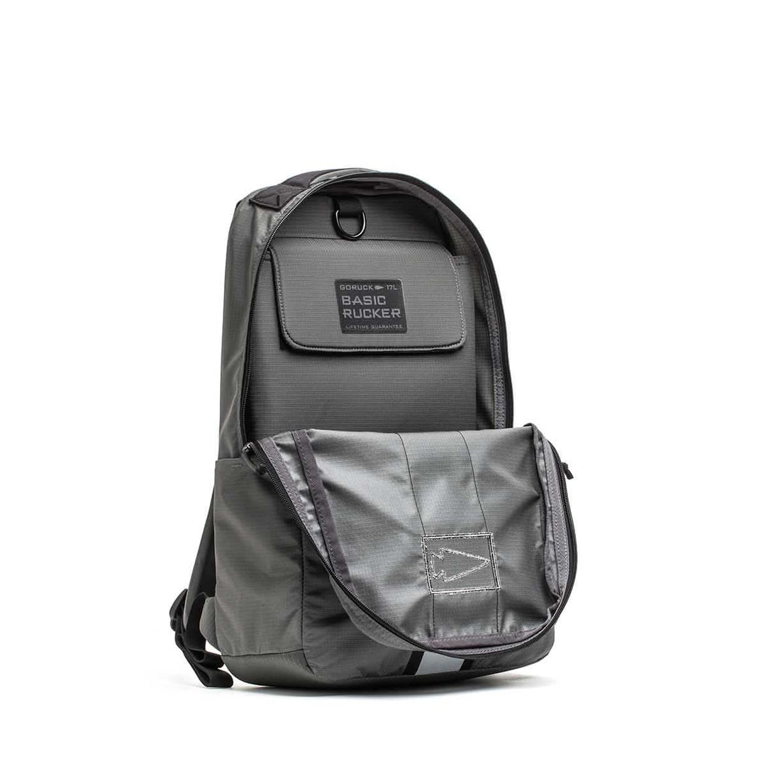 Gray Basic Rucker® backpack with front pocket partially open, revealing the inner compartment and a label reading "GORUCK Basic Rucker.