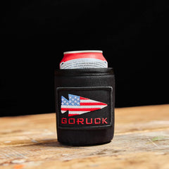 A cold beer rests on a wooden surface in a black GORUCK Ballistic Beer Jacket featuring an American flag arrow patch.