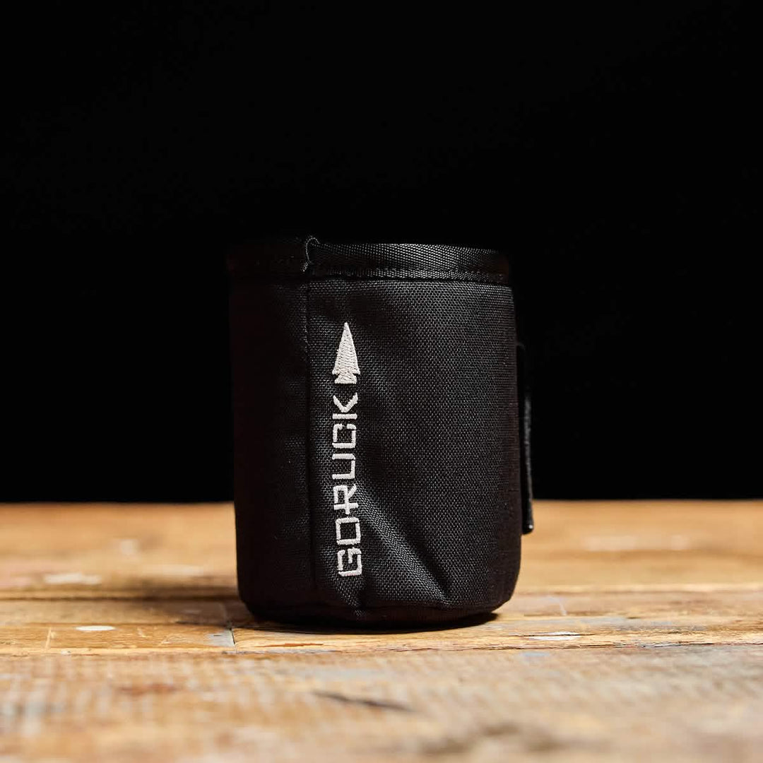 The Ballistic Beer Jacket by GORUCK is a small black pouch made from rugged 1000D CORDURA, featuring a white GORUCK logo, shown on a wooden surface with a black background.
