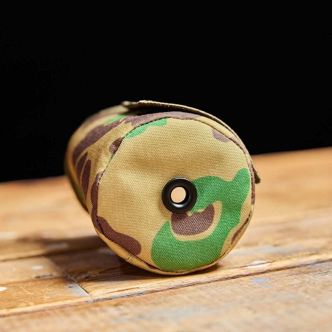 The GORUCK Ballistic Beer Jacket is a camouflage 1000D CORDURA cylindrical pouch with a grommet, perfect as a rugged koozie, shown on a wooden surface.