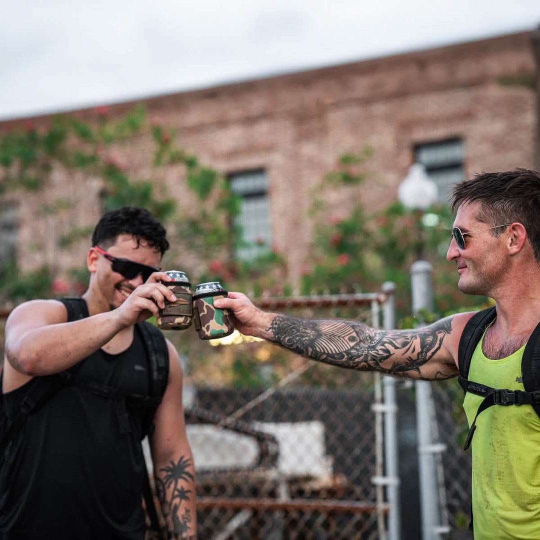 Two men wearing backpacks and sunglasses toast with cold beers kept cool in GORUCK Ballistic Beer Jackets, smiling and enjoying a sunny day outdoors.