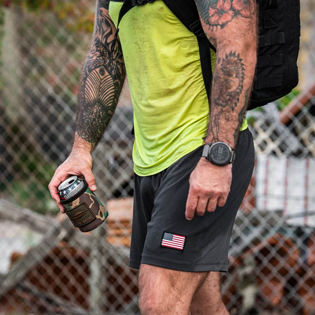 A tattooed person in athletic wear holds a cold beer in the GORUCK Ballistic Beer Jacket, standing by a fence with a U.S. flag patch on their 1000D CORDURA shorts.