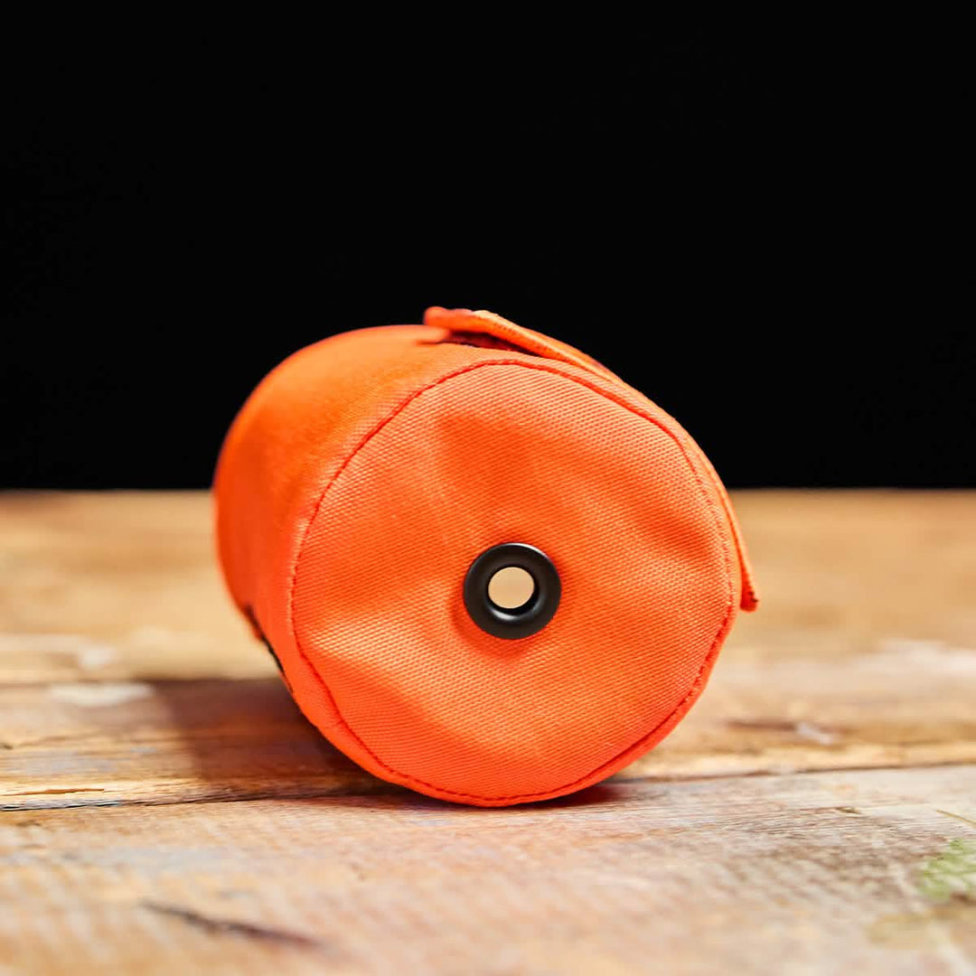 The GORUCK Ballistic Beer Jacket, an orange 1000D CORDURA cylindrical pouch with a black ring, makes the perfect beer koozie and is shown resting on a wooden surface.