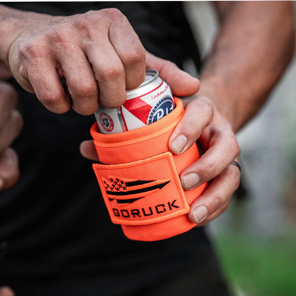 Someone holds a can of cold beer wrapped in a GORUCK Ballistic Beer Jacket, made from rugged 1000D CORDURA and featuring an orange design with a patch.