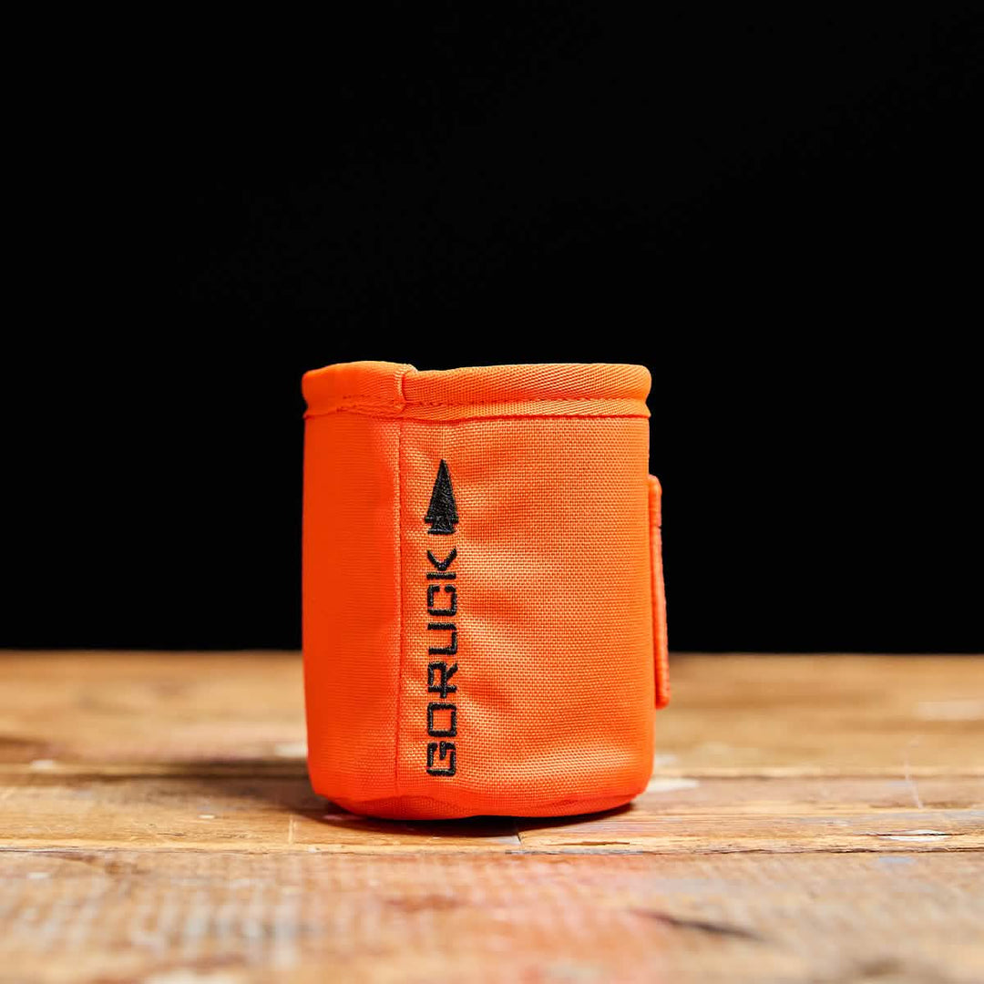 The Ballistic Beer Jacket by GORUCK, made from bright orange 1000D CORDURA fabric with the GORUCK arrow logo, sits on a wooden surface against a black background.
