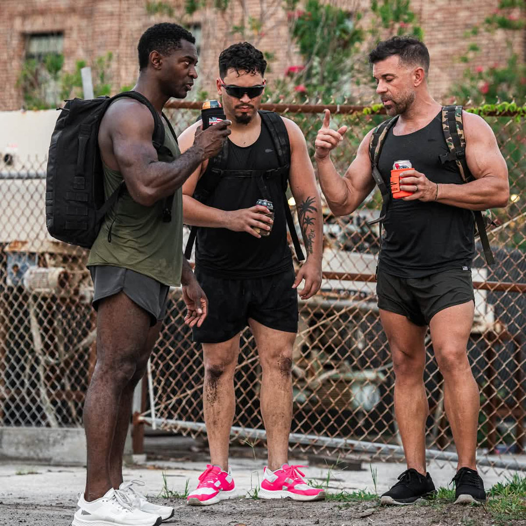 Three men in athletic wear stand outside by a chain-link fence, each holding a cold beer kept chilled in a GORUCK Ballistic Beer Jacket as they chat and unwind.