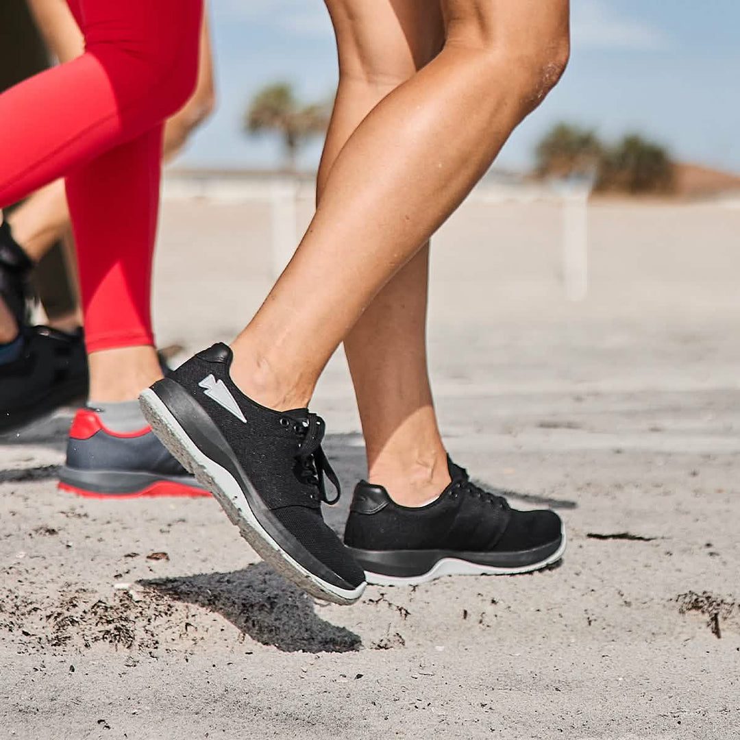 Two people walk on sand in GORUCK Ballistic Trainers - Black + Glacier Grey W / Silver Reflective Spearhead, paired with leggings. Blurred palm trees create a relaxed backdrop.