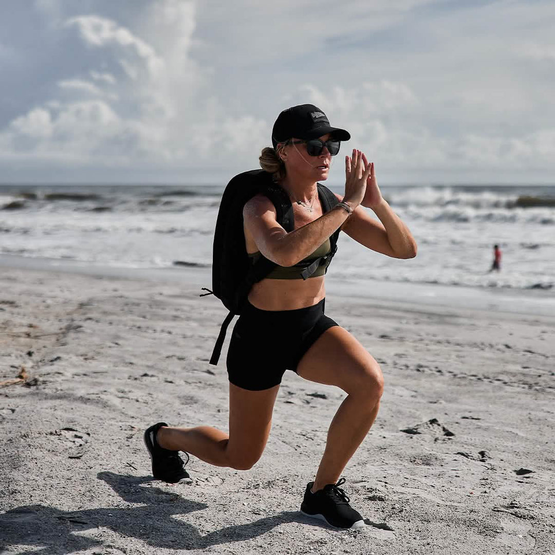 A woman in athletic wear lunges on a sandy beach with ocean waves behind her, showcasing GORUCK Ballistic Trainers in Black + Glacier Grey W / Silver Reflective Spearhead, made with durable CORDURA® Ballistic Nylon.