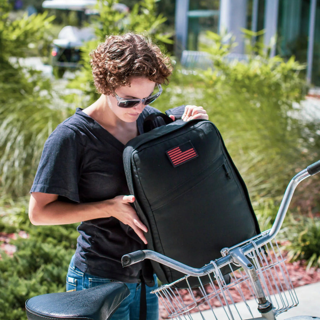Woman placing GORUCK black rucker backpack with USA flag patch into bicycle basket outdoors