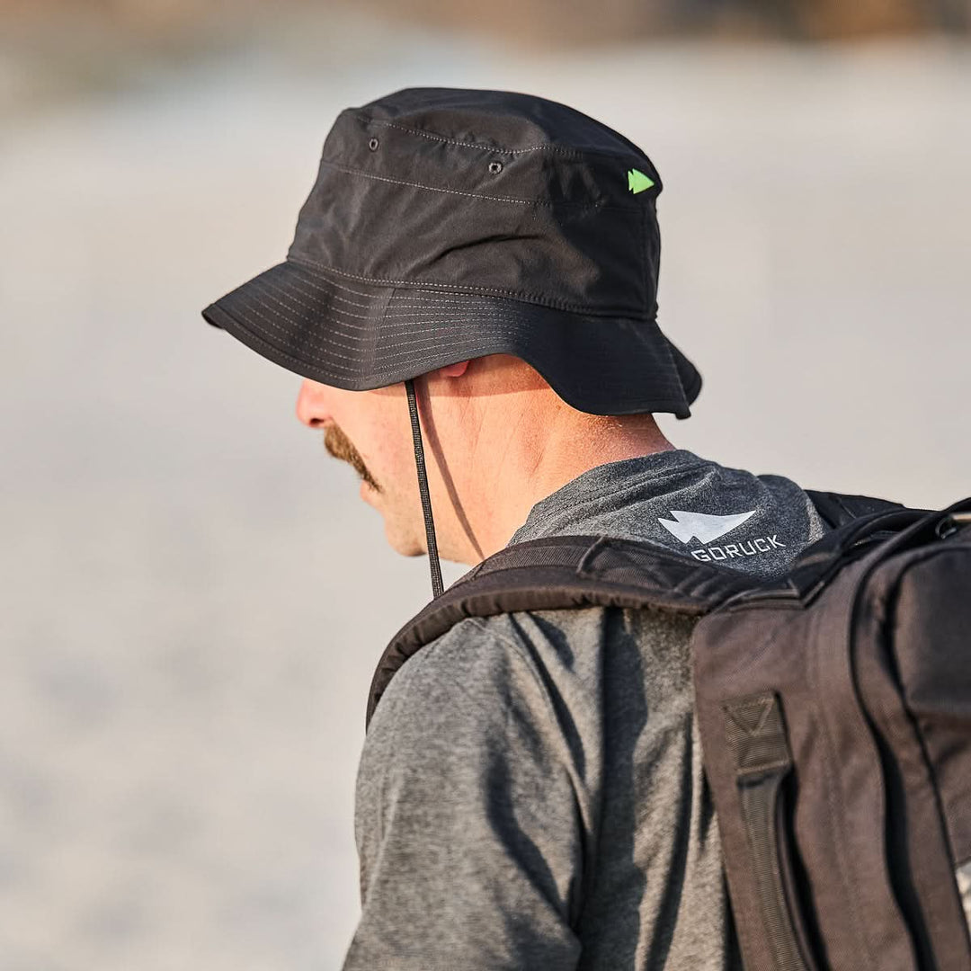 A man with a mustache wearing a black GORUCK Boonie Hat - Slick - ToughDry and a gray shirt carries a black backpack outdoors.
