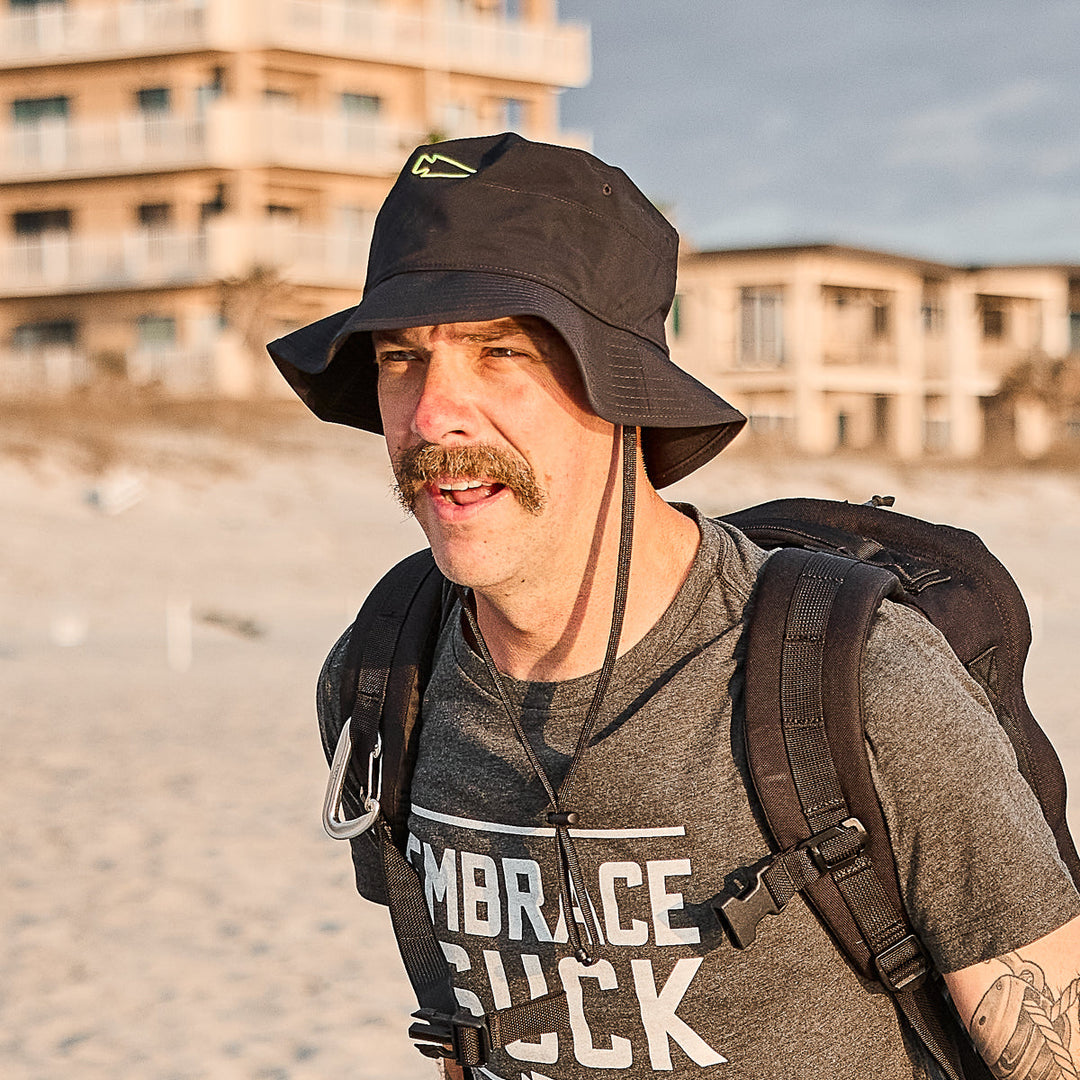 A man with a mustache wears the GORUCK Boonie Hat - Slick - ToughDry and a backpack on a sandy beach, with buildings in the background. The hat offers 50+ UPF for excellent sun protection.