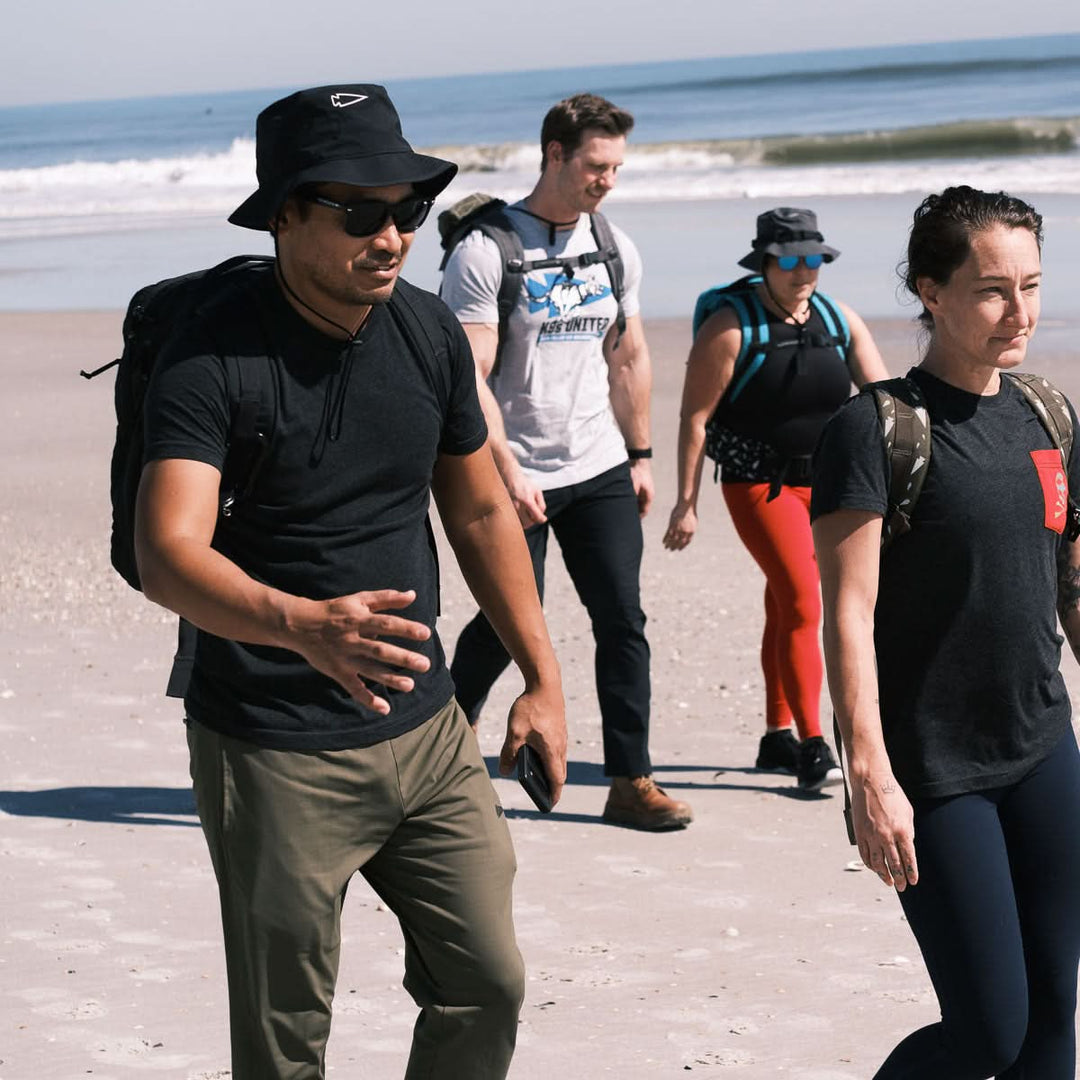 Four people with backpacks, wearing GORUCK Boonie Hat - Slick made from ToughDry® fabric (50+ UPF), walk on a sunny beach near the ocean.