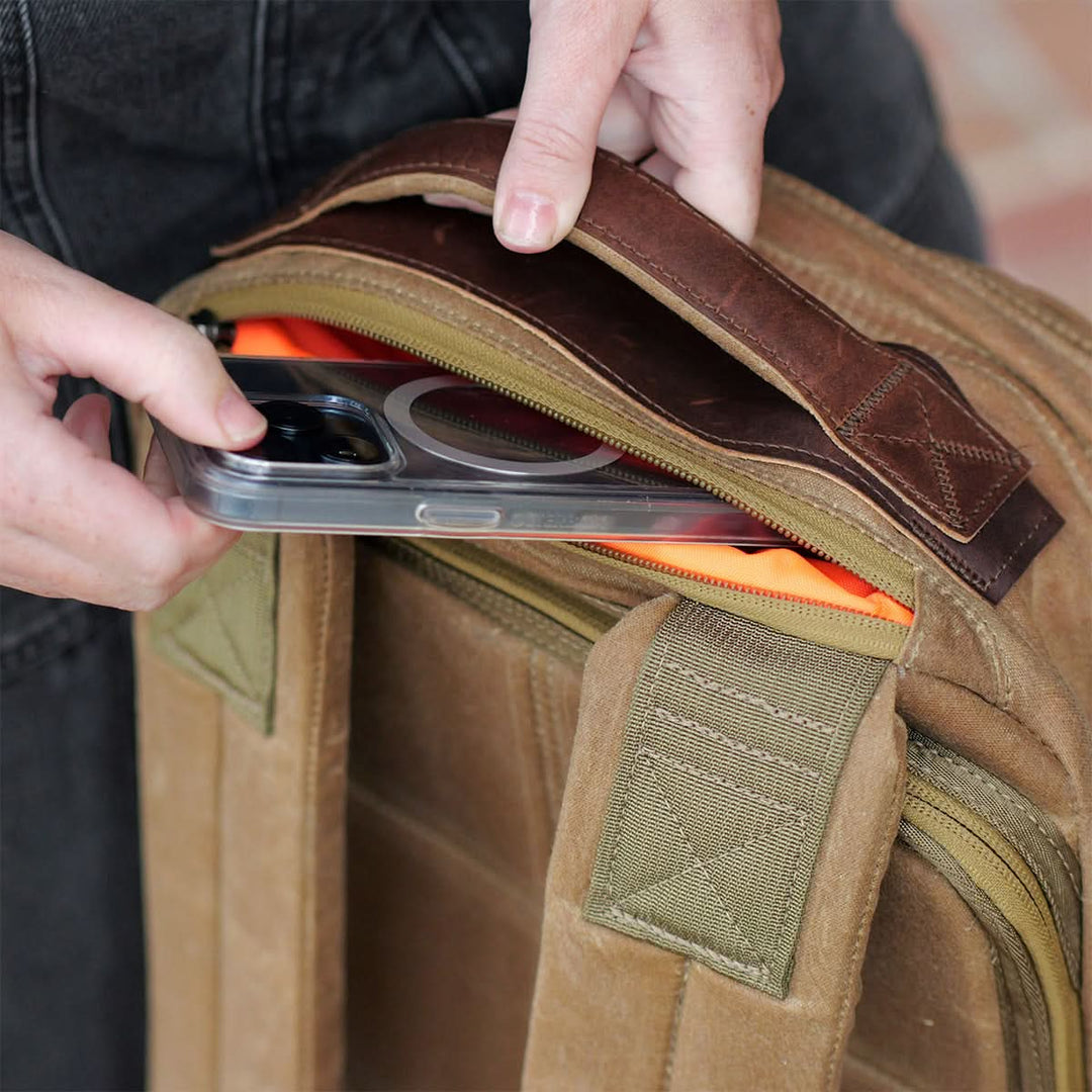Someone is carefully placing a smartphone into a GR1 USA - Heritage rucksack by GORUCK, the brown exterior beautifully complemented by a vibrant orange interior.
