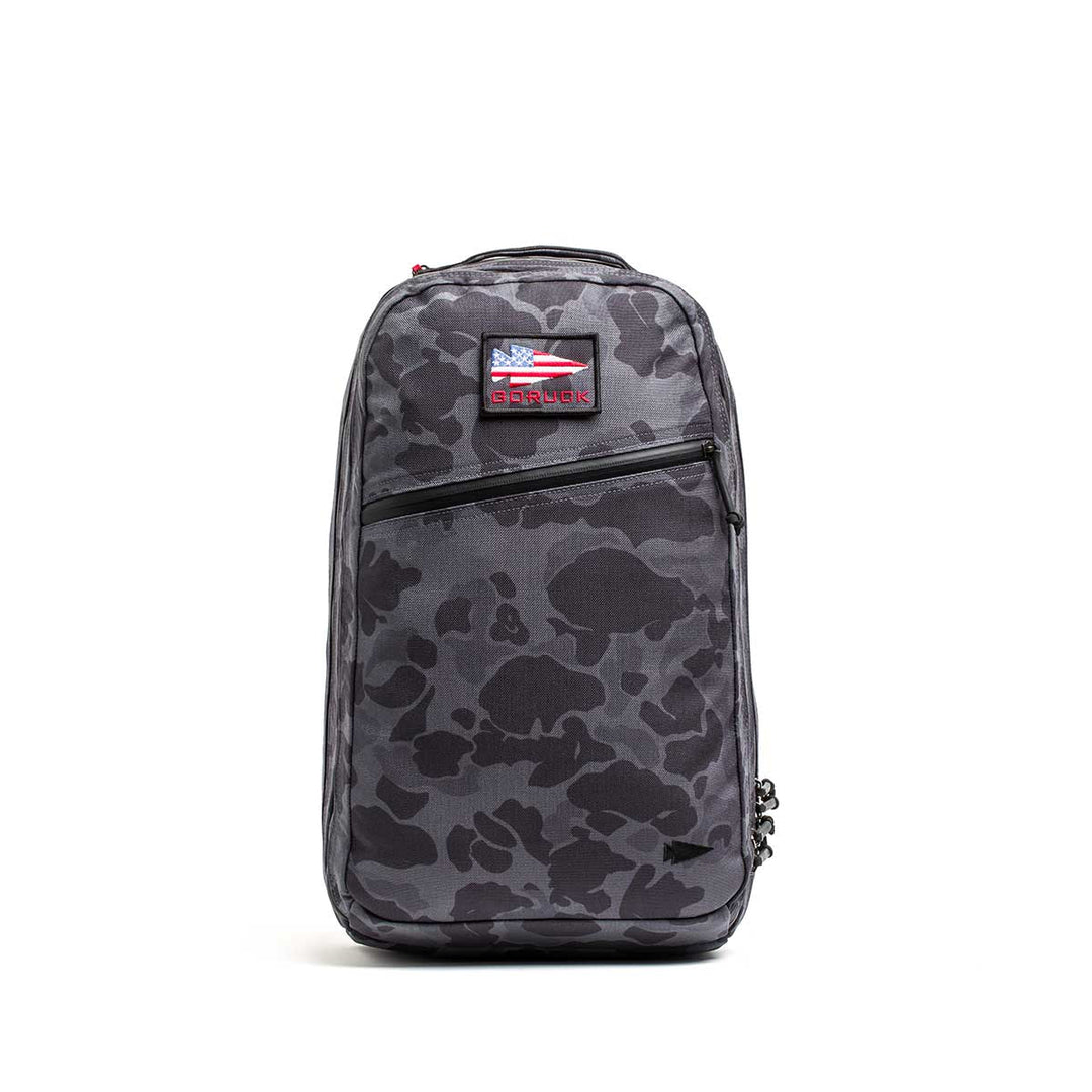 The Double Compartment Bullet Ruck - Ballistic Nylon Cordura - 18L is a gray camo travel backpack with a front zipper pocket, bombproof laptop compartment, and a small USA flag patch near the top.