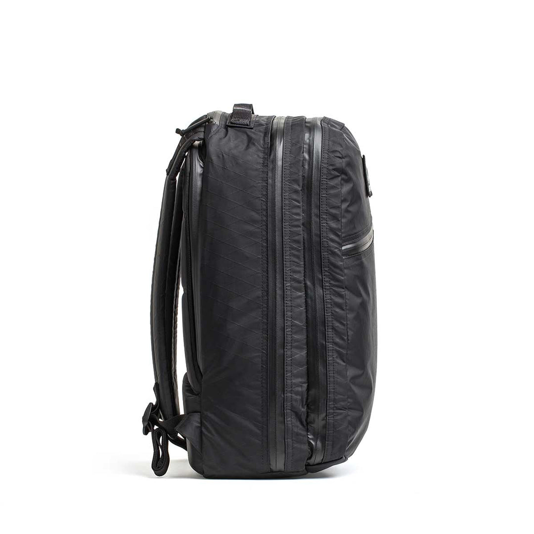 The Double Compartment Bullet Ruck - XPAC, shown from the side against a white background, is a black travel backpack featuring side zippers and an ultra-durable laptop compartment.