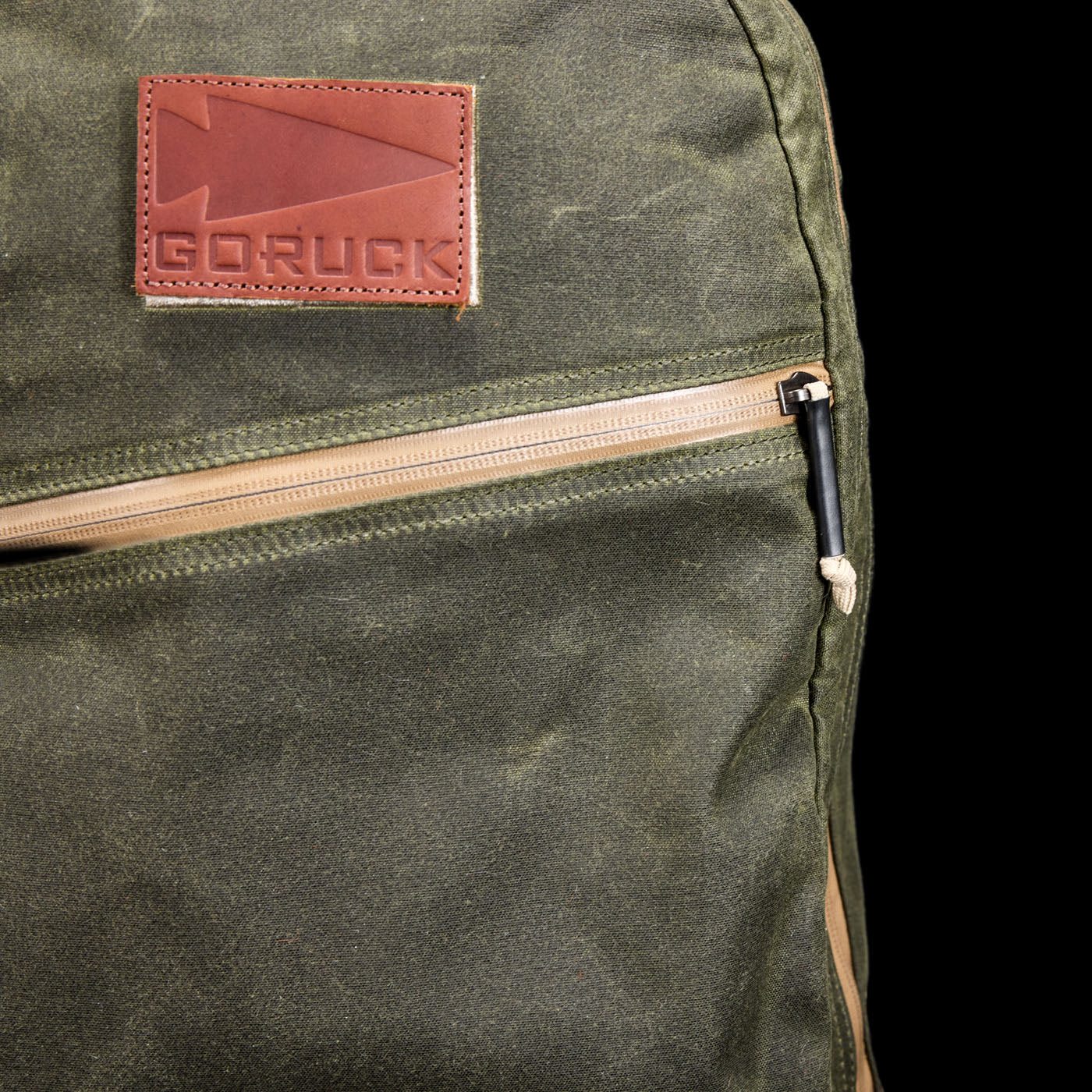 Close-up of a green GORUCK backpack with a brown leather patch and a tan zipper on the front pocket.