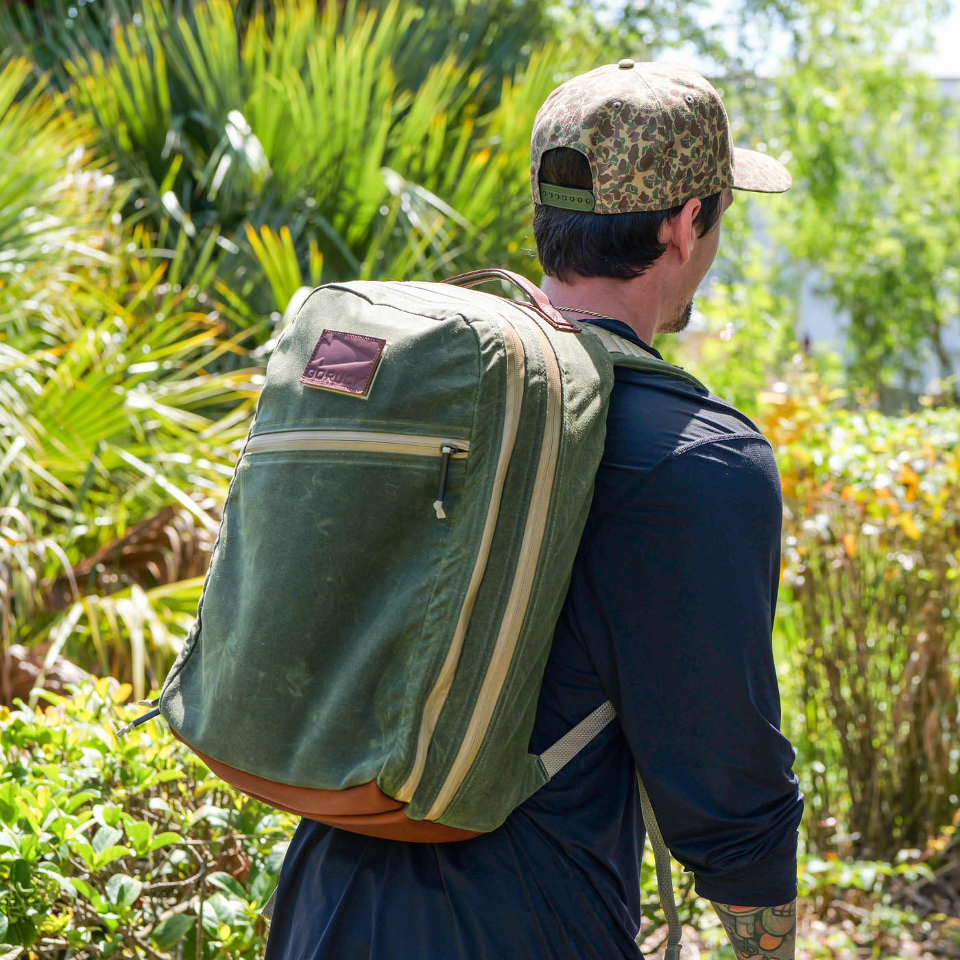 Man wearing a camouflage cap and green backpack walking outdoors near lush green foliage and sunlight.