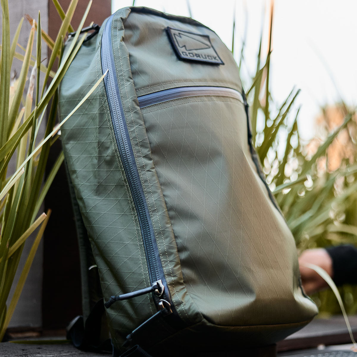 A green backpack with a front zipper pocket, displayed outdoors near tall grass and wooden boards.