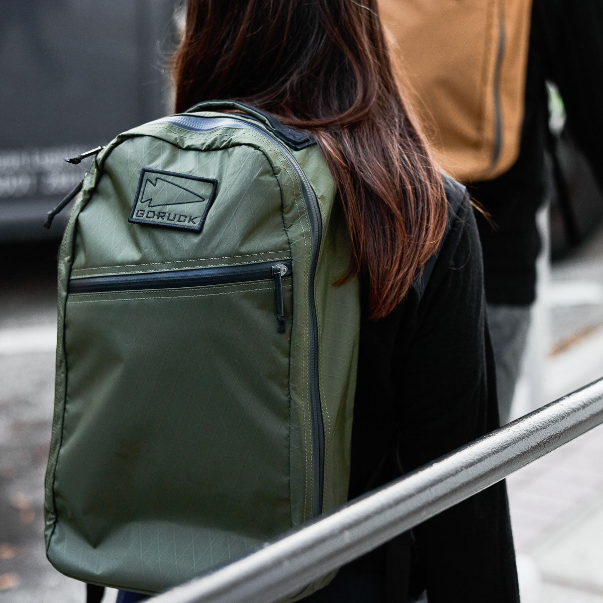 Person wearing a green GORUCK backpack, walking outdoors with another person carrying a tan backpack.
