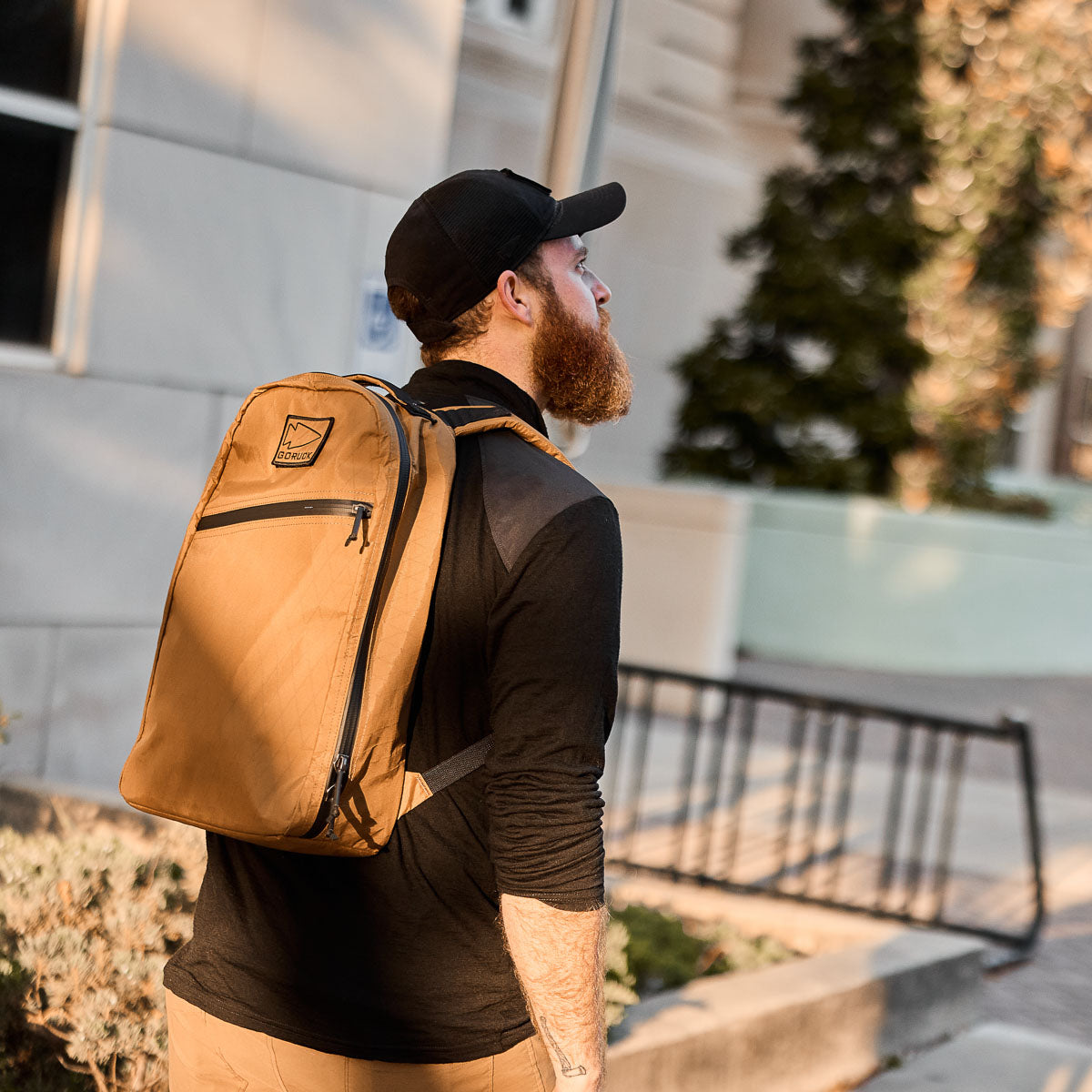 Bearded man in a cap and backpack walks outdoors near a building on a sunny day.