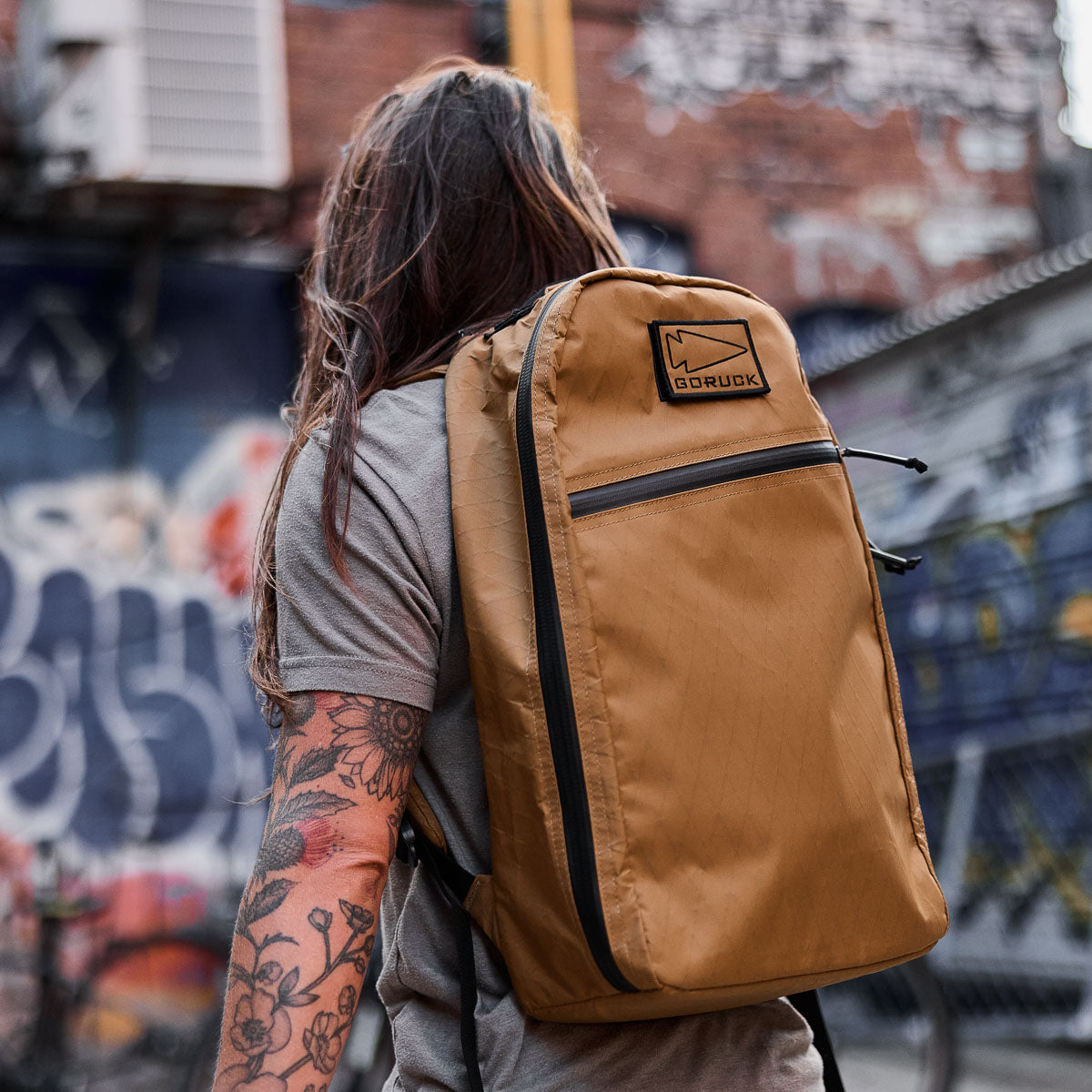 Person with long hair and tattoos wearing a tan GORUCK backpack, standing in front of graffiti wall.