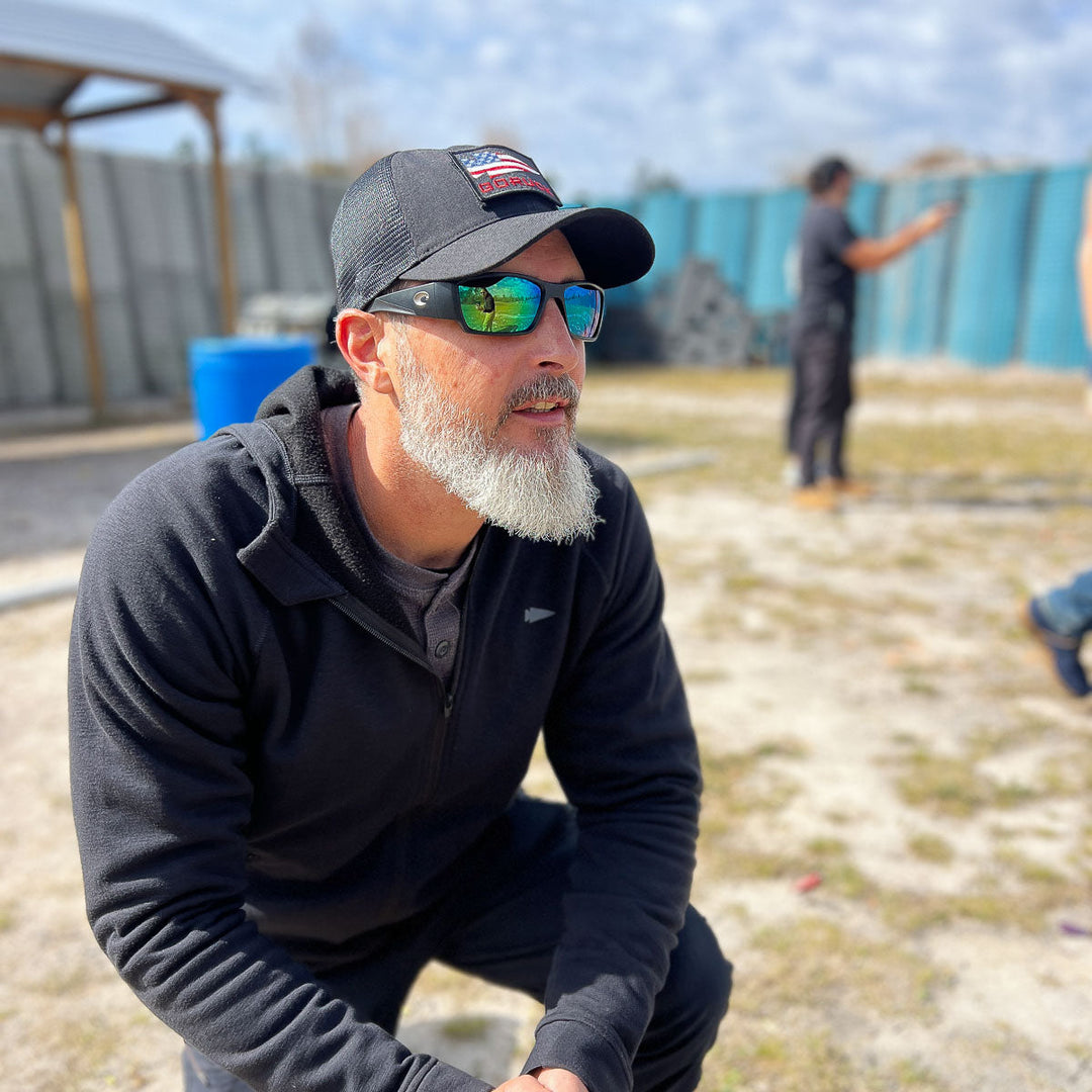 A bearded man in sunglasses and a GORUCK Cadre Hat – CORDURA® kneels outdoors on a sunny day, with blurred figures behind him, radiating the confident presence of a Special Forces Cadre.