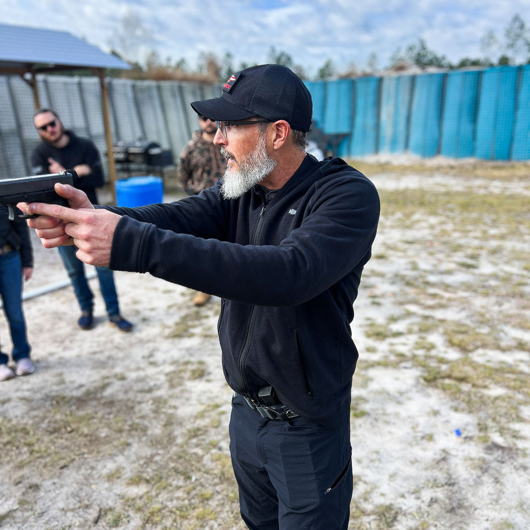 Man wearing a GORUCK Cadre Hat - CORDURA® and sunglasses aims a handgun at an outdoor shooting range, evoking the style of Special Forces Cadre.