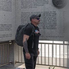 A man in sunglasses and a ToughDry® fabric backpack stands reading a wall inscribed with text, while his GORUCK Performance TAC Hat - TOUGHDRY shields him from the sun.