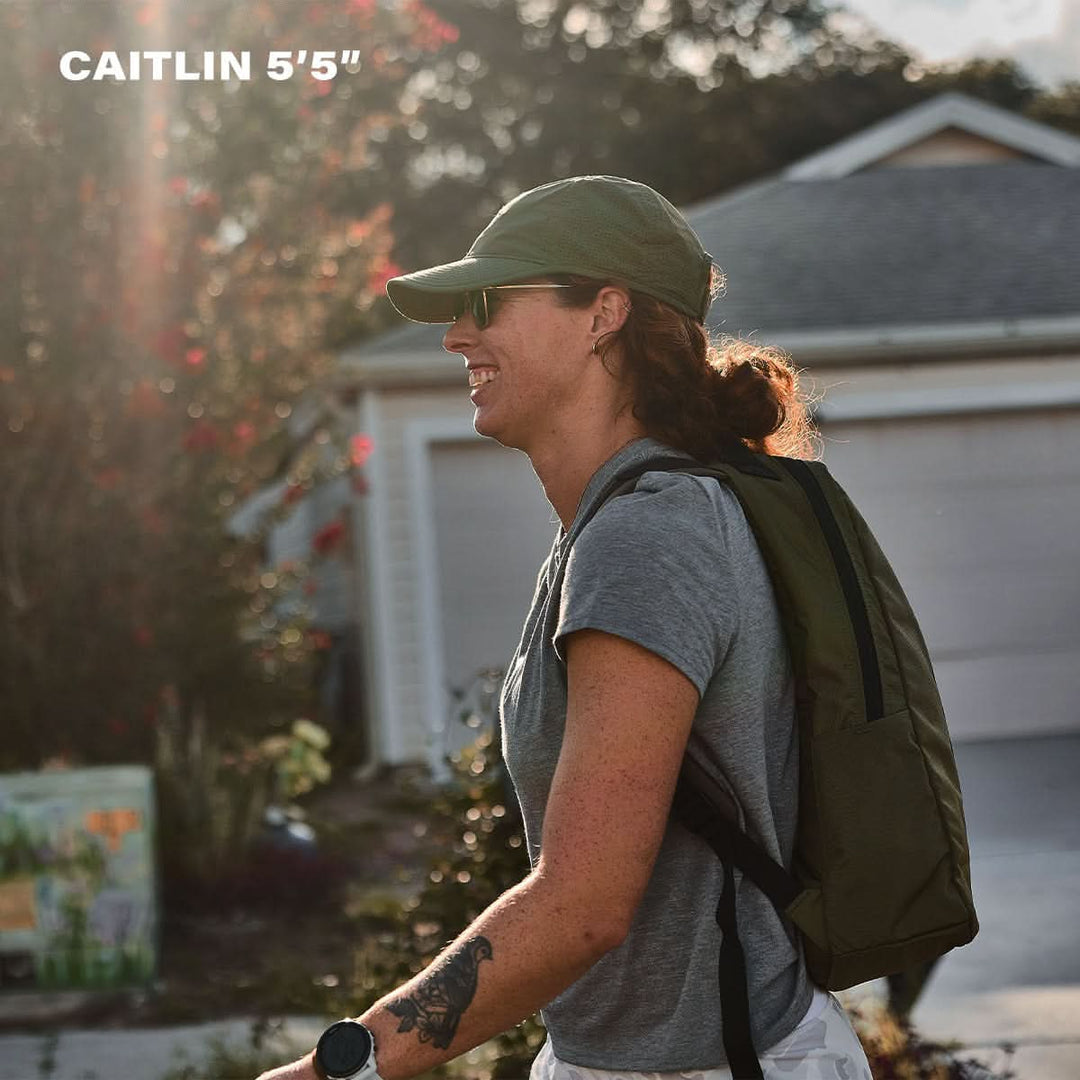 Woman with sunglasses, hat, and the Basic Rucker® backpack smiles outdoors; text reads "CAITLIN 5'5"" in upper left corner.