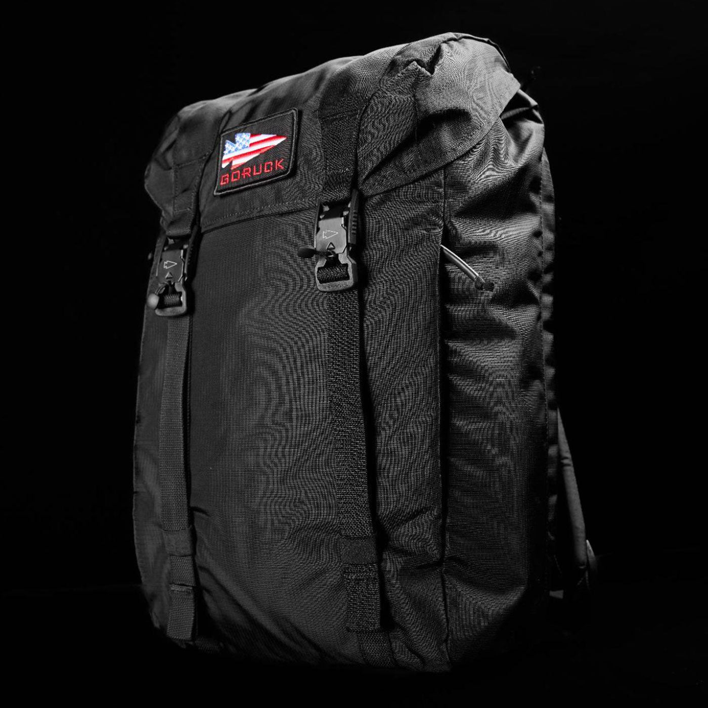 Black backpack with buckled flap and an American flag patch on the front, against a black background.