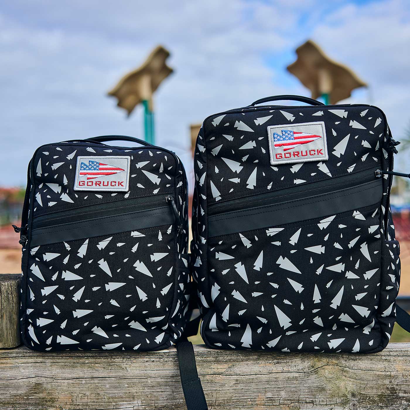 Two black GORUCK backpacks with white patterns and American flag patches on a wooden fence outdoors.