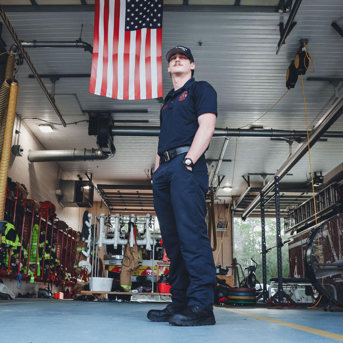 A firefighter stands in a fire station under an American flag, wearing a uniform and cap.